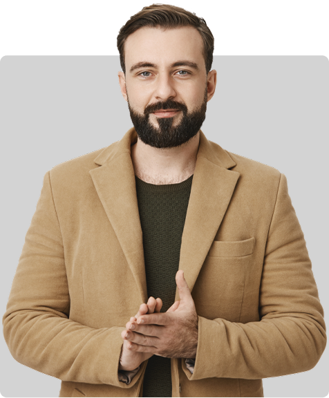 A man with a light brown/beige jacket, dark green shirt, and a beard is looking directly at the camera. His hands are clasped in front of him.